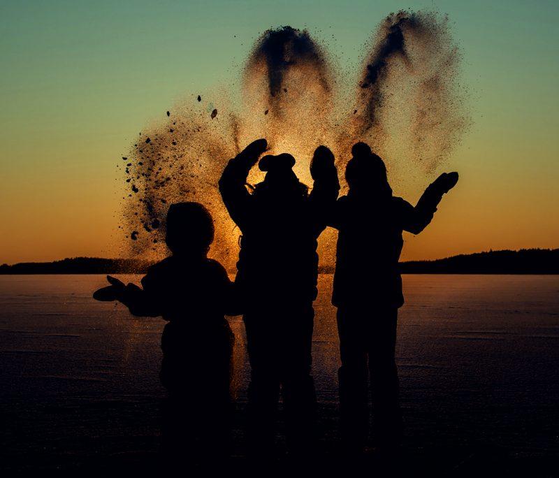 kolme lasta heittää lunta ilmaan ilta-auringossa