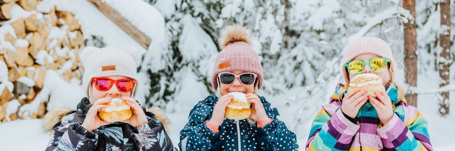 kolme lasta istuu lumihangessa aurinkolasit päässään ja syövät laskiaispullia