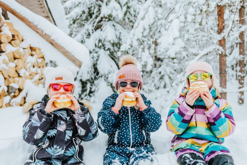 kolme lasta istuu lumihangessa ja syö aurinkolasit päässään isoja laskiaispullia